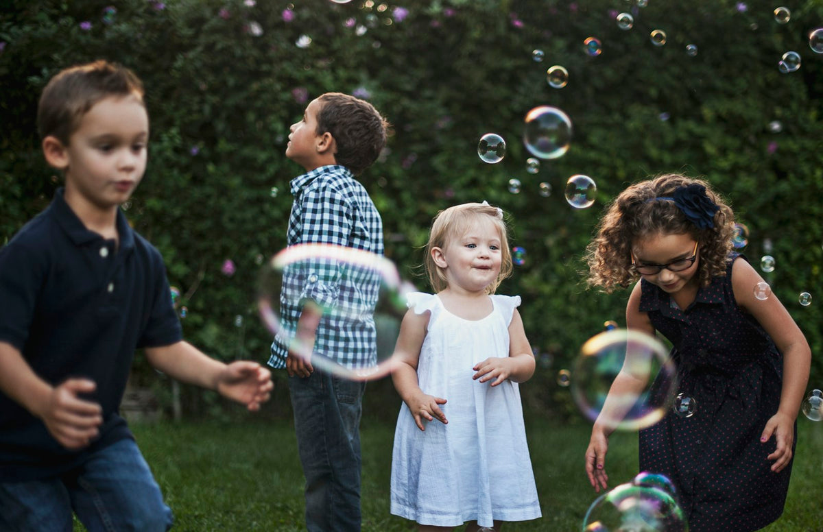 Kids chasing bubbles in a backyard, a toddler in a white dress in the middle